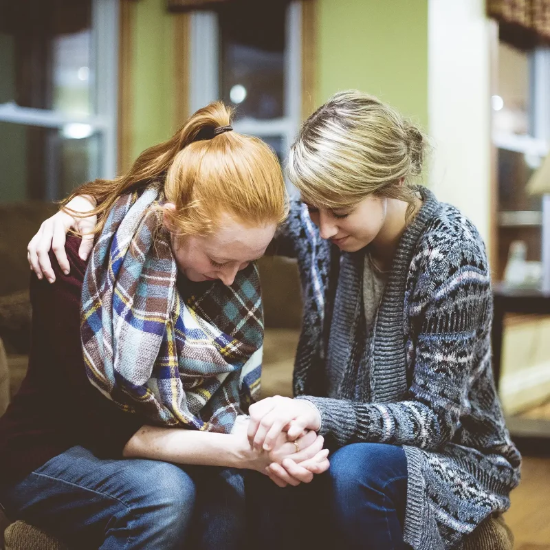 Two young female friends sitting on the couch and praying together