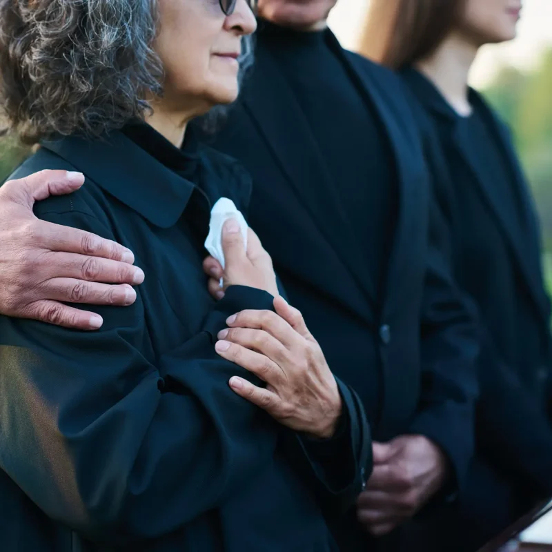 Hand of mature man on shoulder of his wife or sister with handkerchief lamenting passes away relative or family member during funeral service