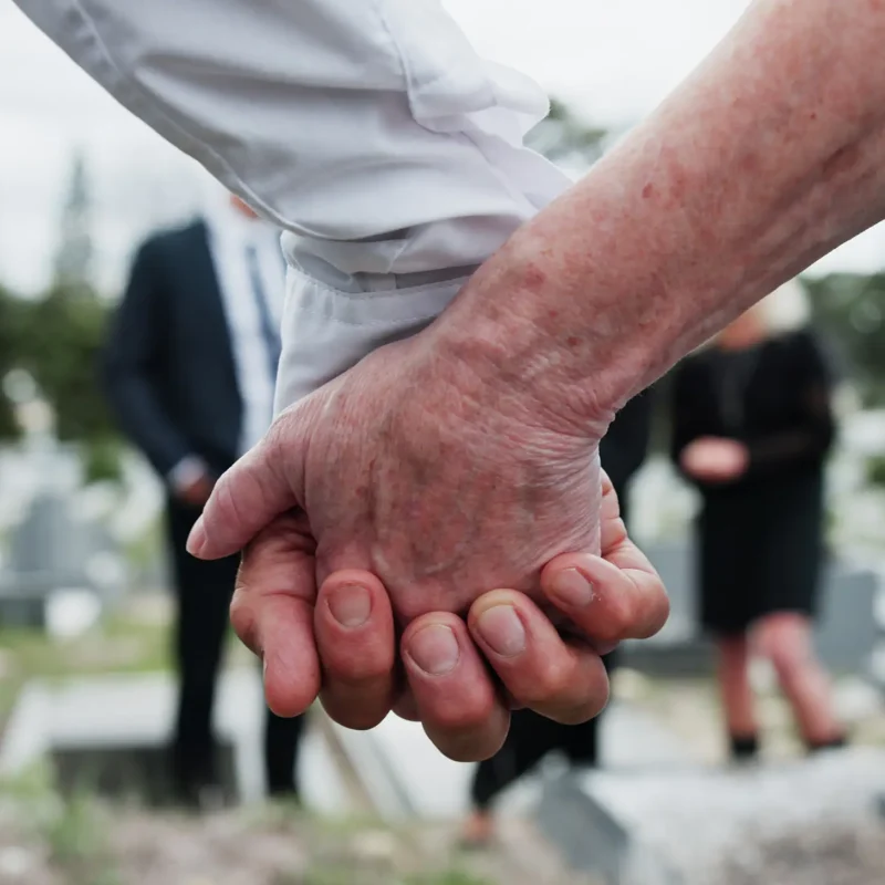 Funeral, people and holding hands in graveyard with grief, mourning and empathy for burial ceremony. Family, death and senior person at cemetery outdoor with memorial service, bereavement or support.