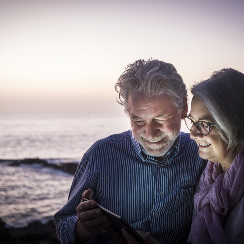 Happy retired couple having fun sitting on the cliffs, dusk light looking at the same tablet. Happy retirement concept. Sea in the background