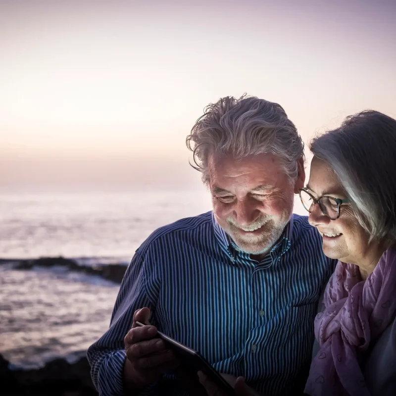 Happy retired couple having fun sitting on the cliffs, dusk light looking at the same tablet. Happy retirement concept. Sea in the background